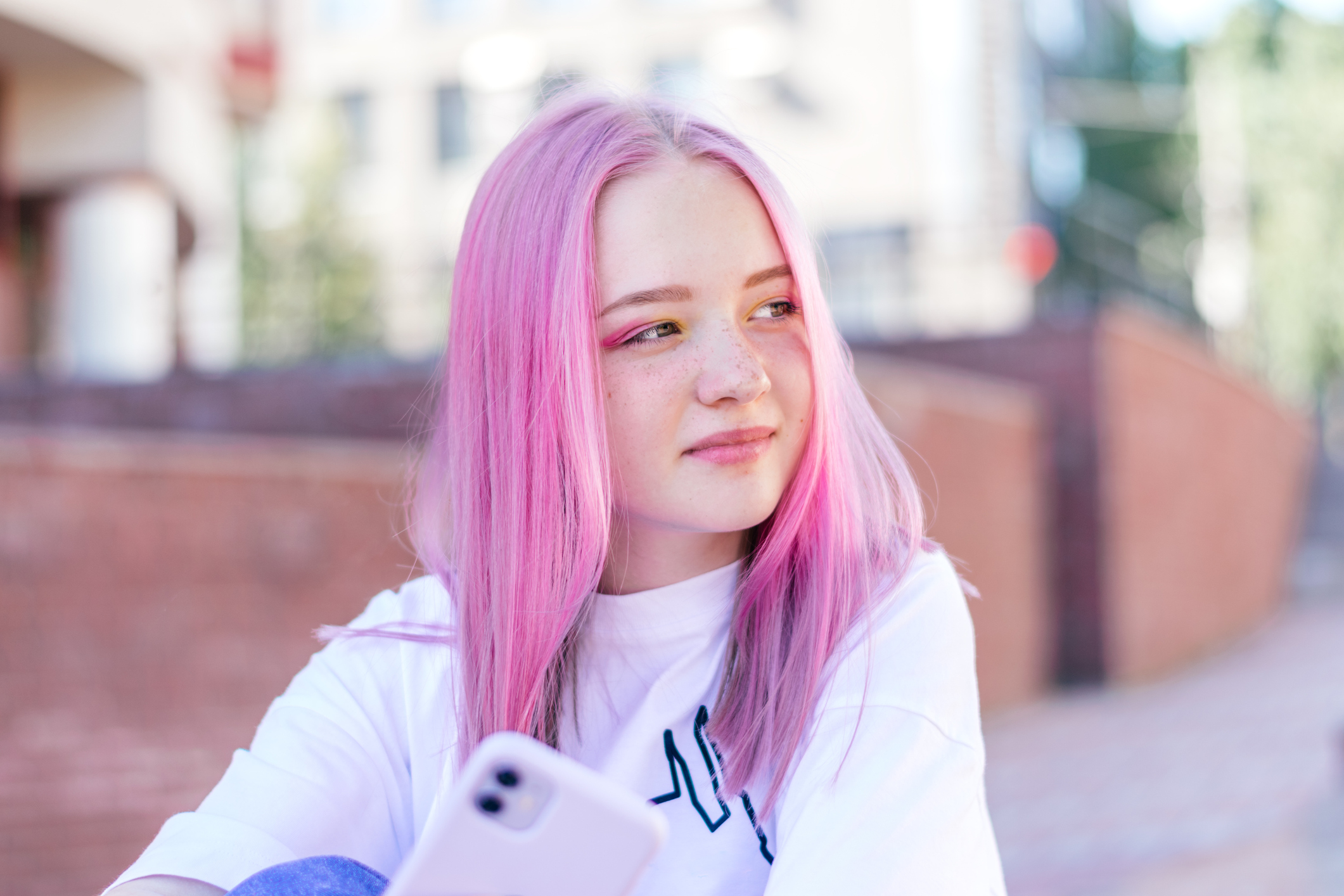 Positive pink-haired teenage hipster girl in a white T-shirt is using a smartphone on a summer day on the street of the city.Summer concept.Generation Z style.Social media concept
